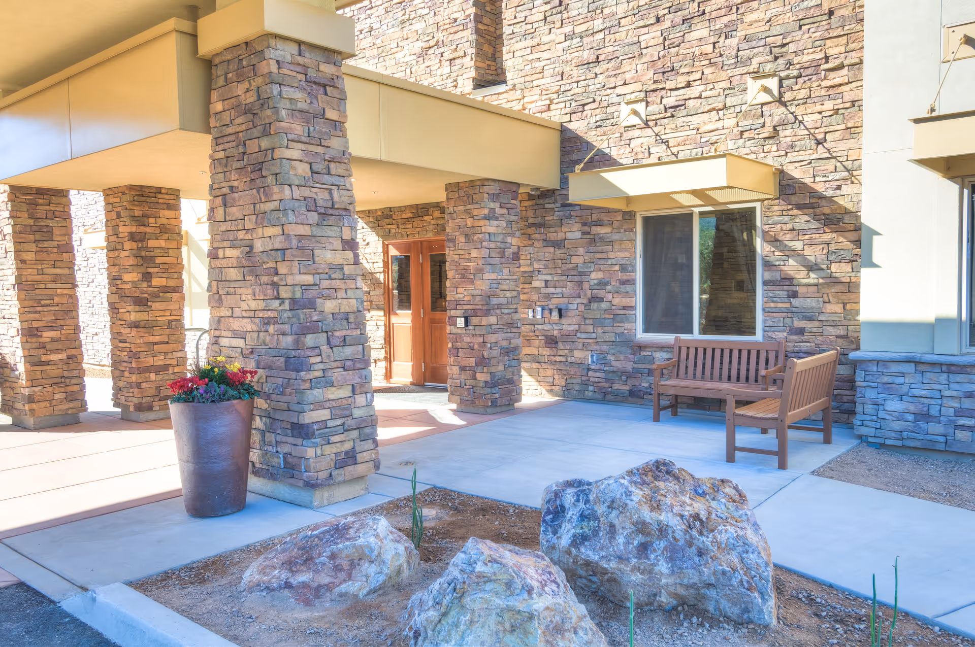 Stone-columned building entrance with a wooden bench, potted flowers, and decorative rocks in the foreground.