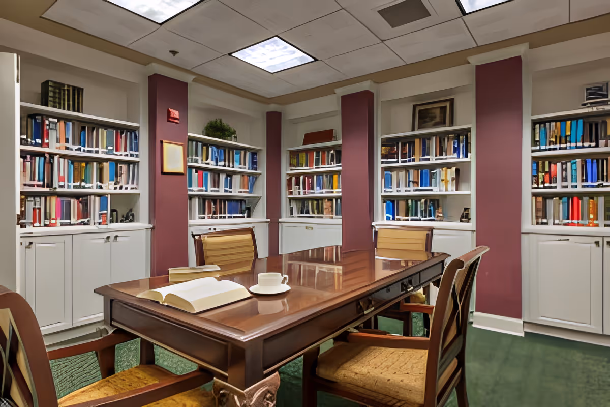 A cozy library room with a wooden table and four chairs. The table has an open book and a cup on it. Surrounding the room are built-in white bookshelves filled with colorful books. The walls have maroon columns, and the ceiling has recessed lighting panels.