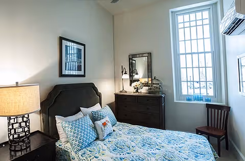 Bedroom with a blue-and-white patterned bed, dark wood nightstands and dresser, a tall window, and a wall-mounted air conditioner.