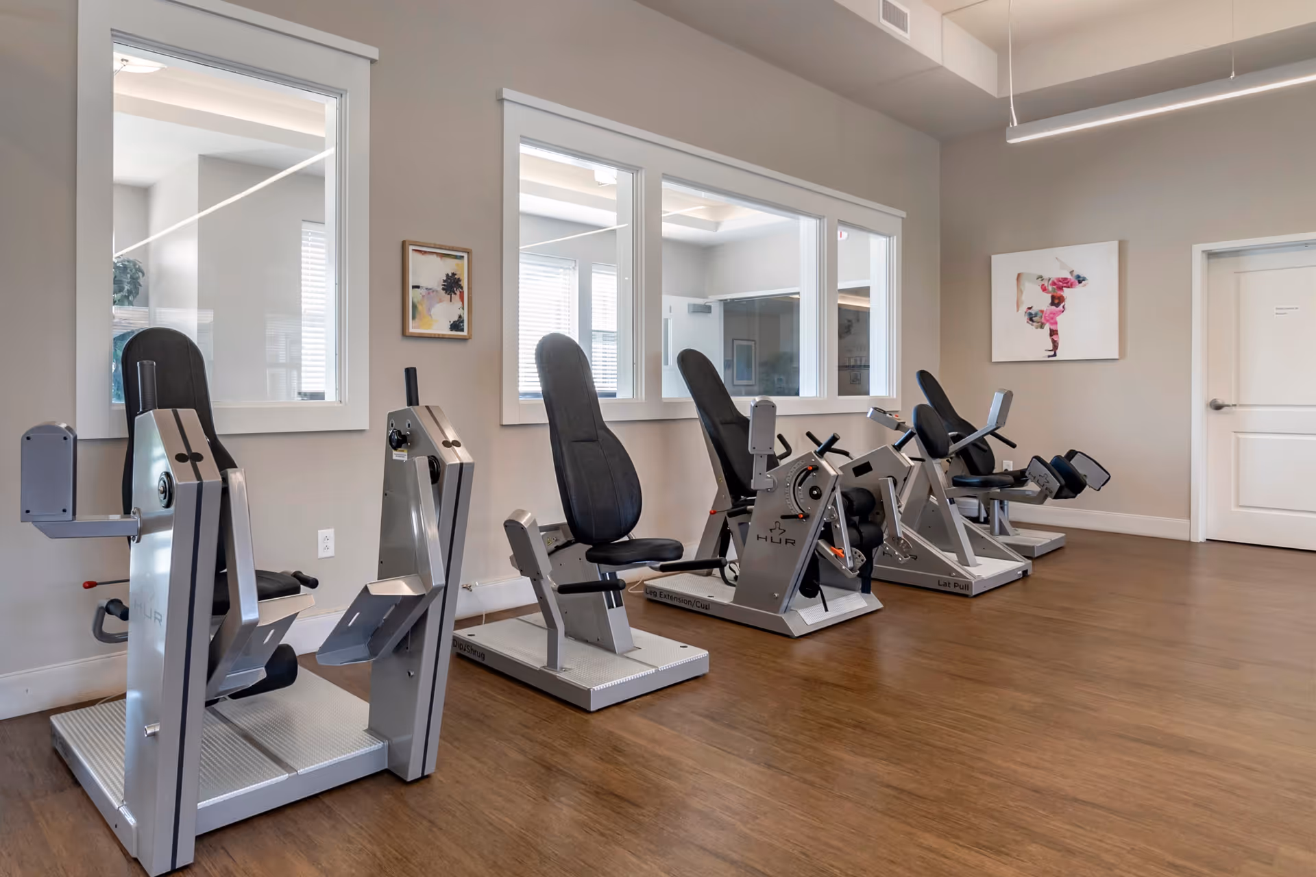 A senior living community exercise room with four seated strength training machines lined up against a wall with large windows. The room has wood flooring, neutral-colored walls, and a piece of artwork depicting a person doing a handstand on the wall near a closed door.