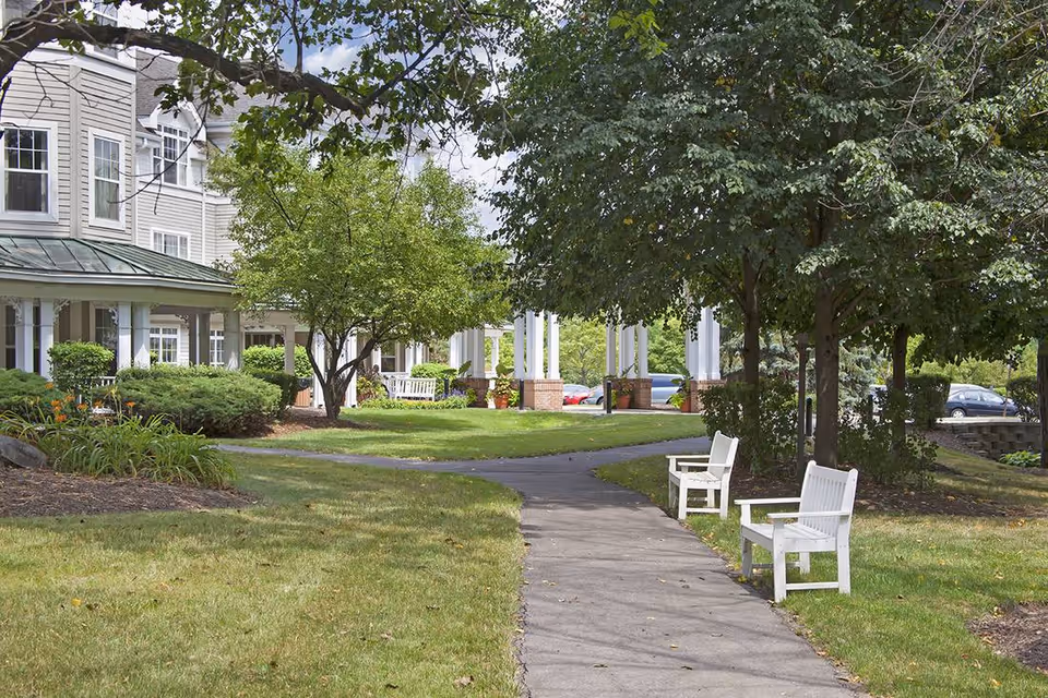 A paved walkway with white benches leading to the front entrance of a senior living building surrounded by trees and landscaping.