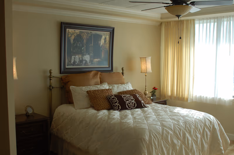 A cozy bedroom with a neatly made bed featuring multiple pillows in shades of brown and cream. There is a framed painting above the bed, a nightstand with a lamp and a small clock on the left side, and another nightstand with a lamp and a small red flower on the right side. Light yellow curtains cover a large window, allowing soft natural light to fill the room. A ceiling fan is visible above the bed.
