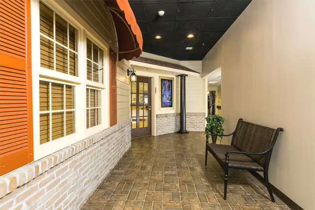 Indoor hallway styled like a streetscape with a bench, storefront-style window and door, brick half-wall, awning, and potted plant.