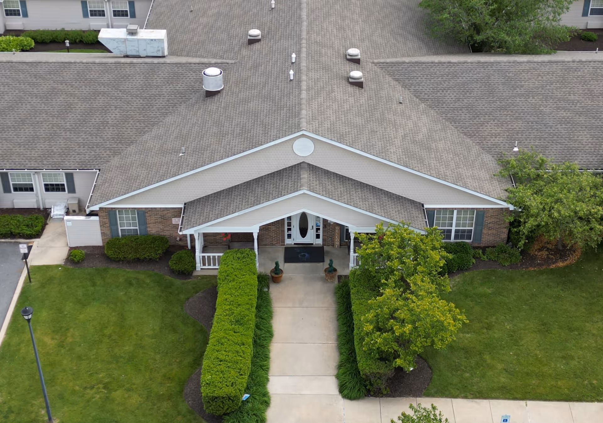 Aerial view of the front entrance of a single-story residential building with a gabled roof, surrounded by green lawns, bushes, and trees. A concrete walkway leads to the covered porch entrance with white pillars and a glass front door.