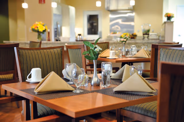 Well-lit dining room with neatly set wooden tables, folded napkins, glassware, and small floral centerpieces.