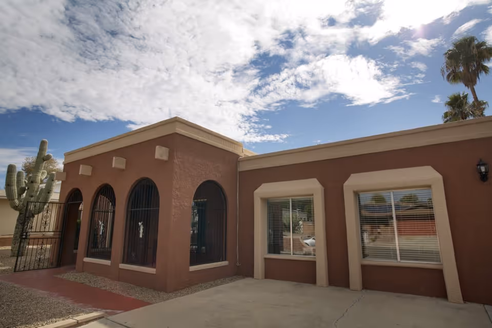Single-story stucco building with arched gated windows, rectangular windows, and desert landscaping under a partly cloudy sky.