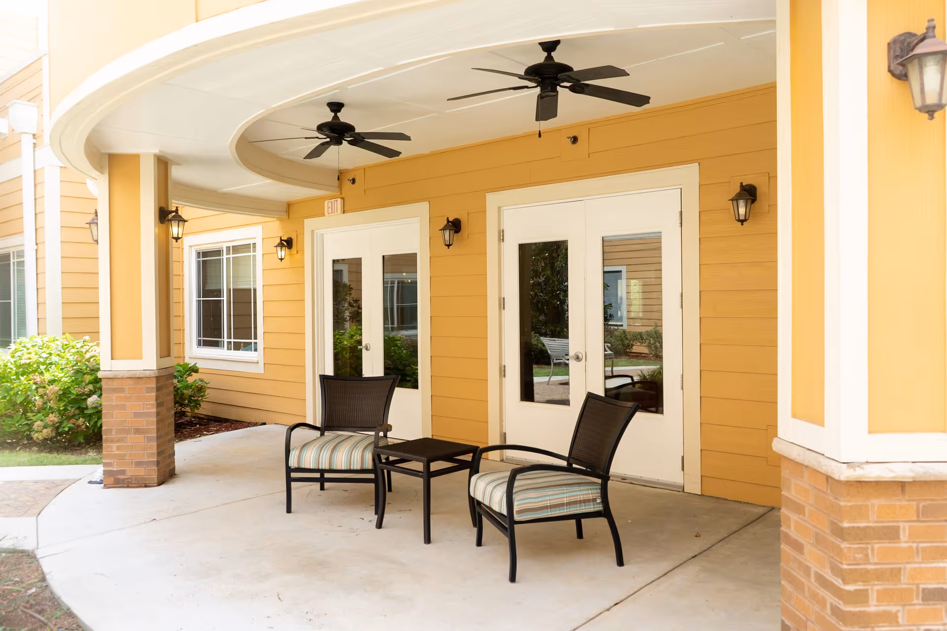 Covered outdoor patio area with two black chairs with striped cushions and a small black table between them. The patio has a yellow exterior wall with two white-framed glass doors, wall-mounted lantern-style lights, two ceiling fans, and brick pillars. Green shrubs and a window are visible in the background.