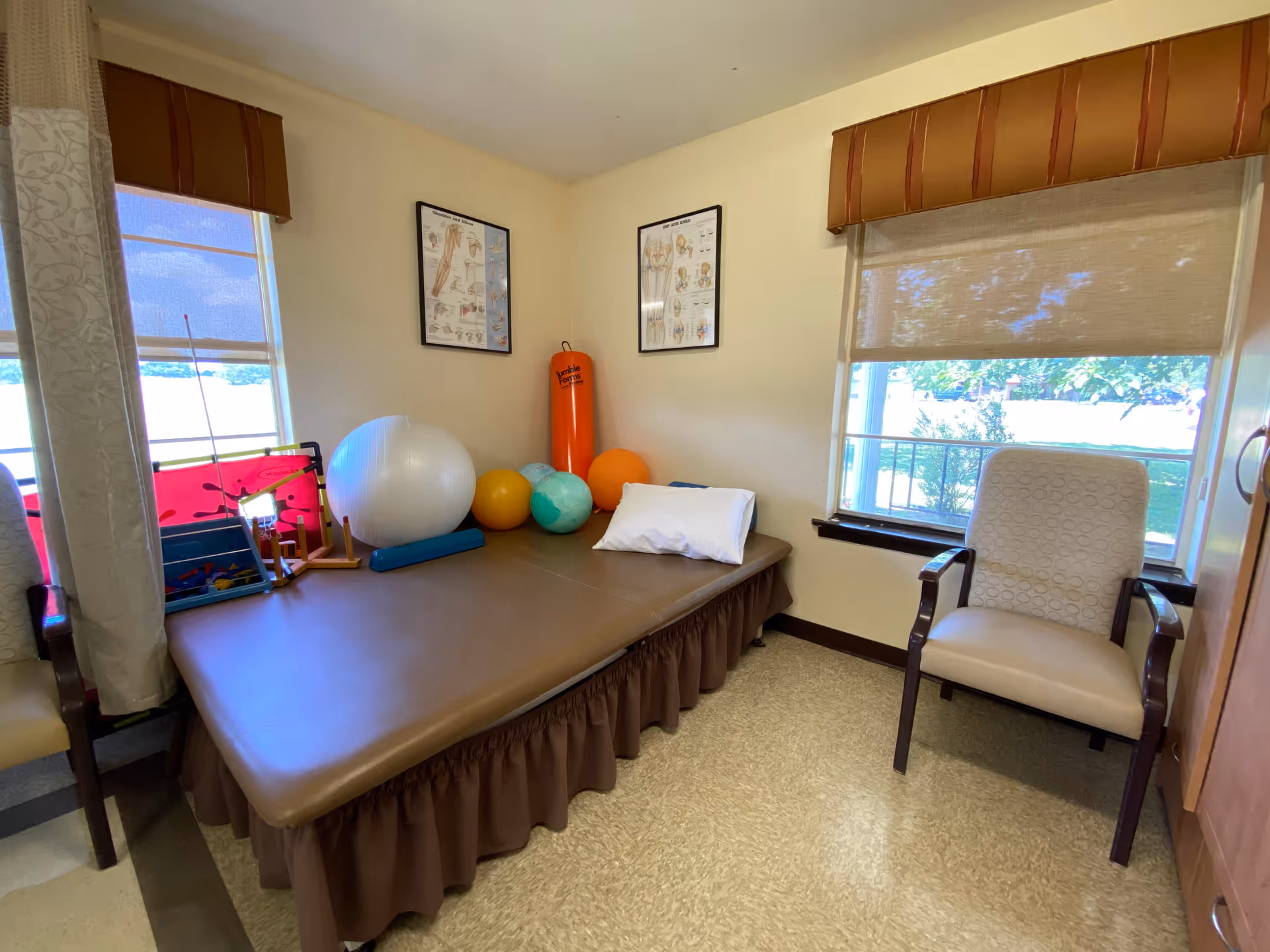 Small therapy room with a padded treatment table holding exercise balls and a pillow, two windows, an armchair, and anatomy charts on the wall.