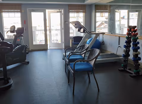 A fitness room with exercise equipment including a treadmill, stationary bike, and a rack of dumbbells. Two chairs with blue cushions are placed in the center of the room. Large windows and glass doors let in natural light, and a mirrored wall reflects the room.