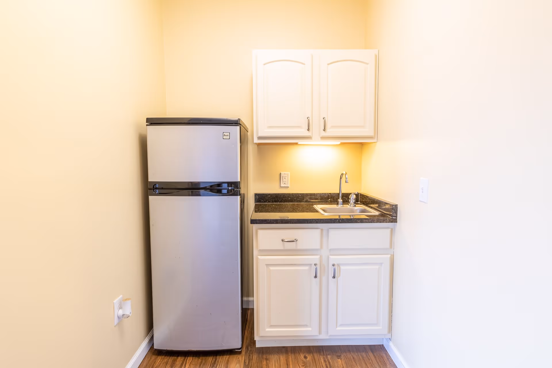 Small kitchenette area with a stainless steel refrigerator on the left, white cabinets above and below a black countertop, and a sink with a faucet. The walls are painted light yellow and the floor is wooden.