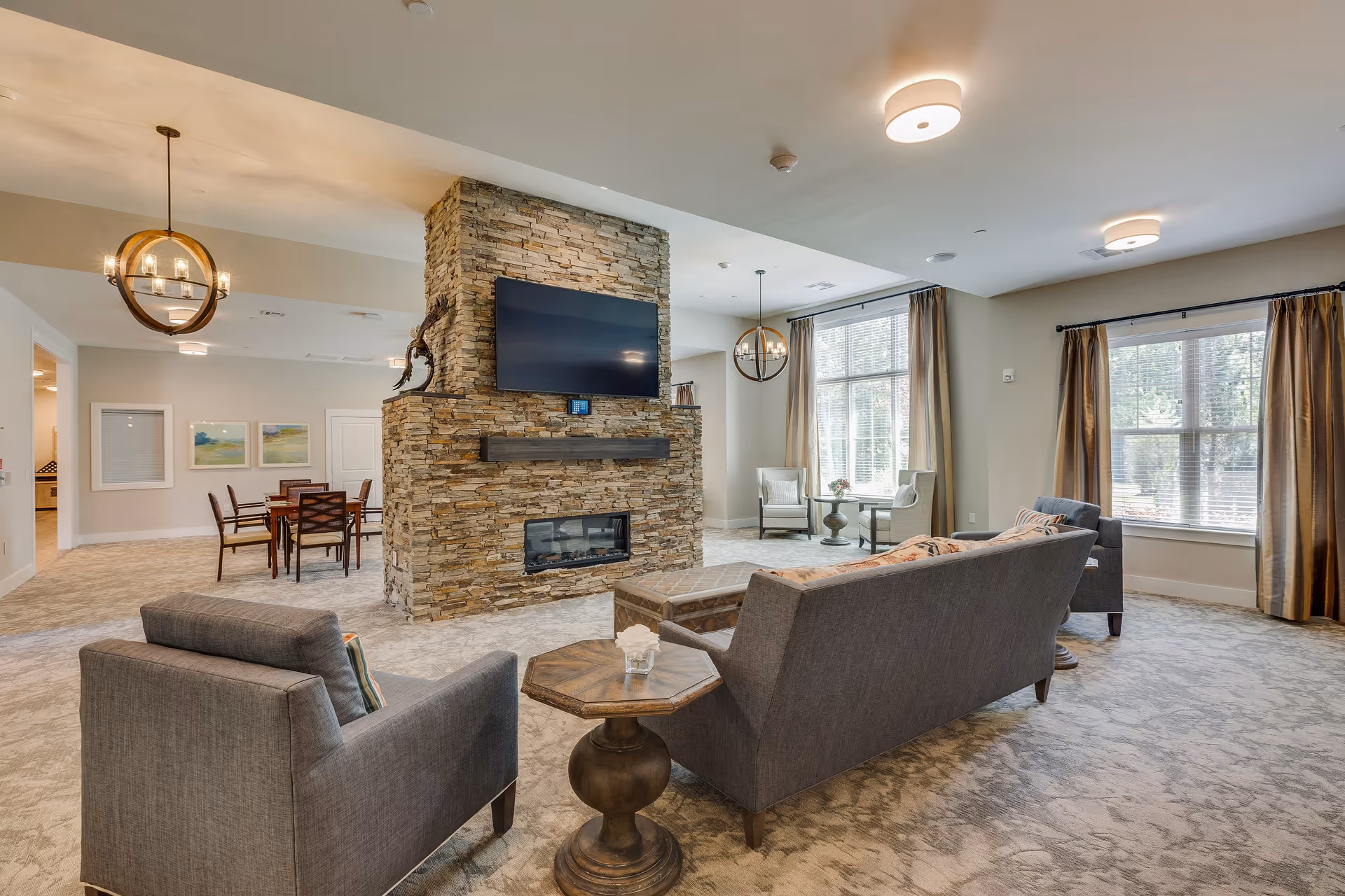A spacious living room area with a stone fireplace and a mounted flat-screen TV. The room features two gray sofas, a wooden side table, large windows with beige curtains, and a dining table with chairs in the background. The space is well-lit with ceiling lights and pendant lamps.
