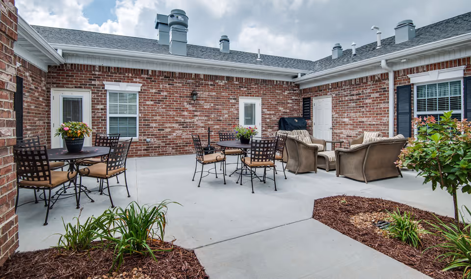 Outdoor patio area with brick walls, featuring two round tables with metal chairs and cushions, a seating area with cushioned wicker furniture, potted plants on the tables, and landscaped garden beds with plants and mulch.