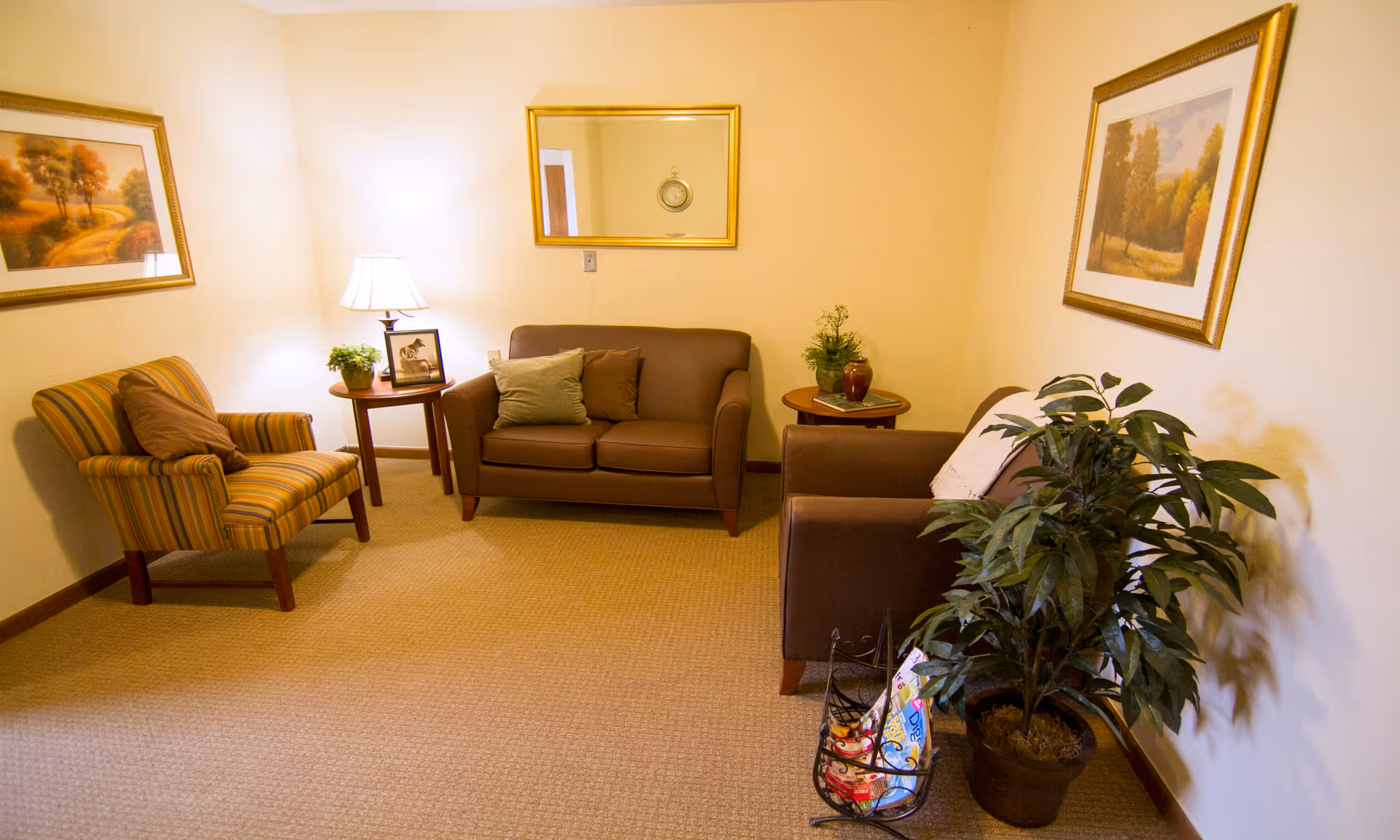 A cozy living room with a brown loveseat and matching armchair, a striped armchair, two wooden side tables with plants and a lamp, framed landscape paintings on the walls, a large mirror, and a potted plant next to a magazine rack.
