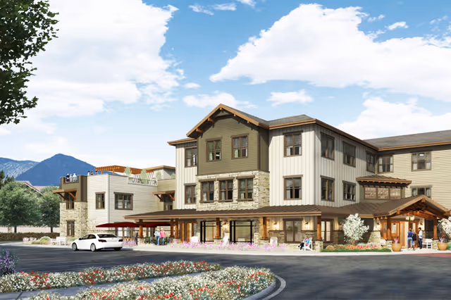 Exterior view of a three-story senior living facility building with a mix of stone and siding facade, surrounded by landscaped flower beds and a curved driveway. People are seen near the entrance and on a rooftop terrace. Mountains and a partly cloudy sky are visible in the background.