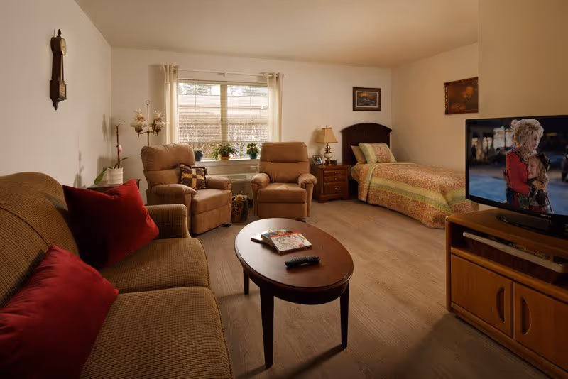 Cozy studio room with a bed, sofa and two recliners arranged around a coffee table and television beneath a window.