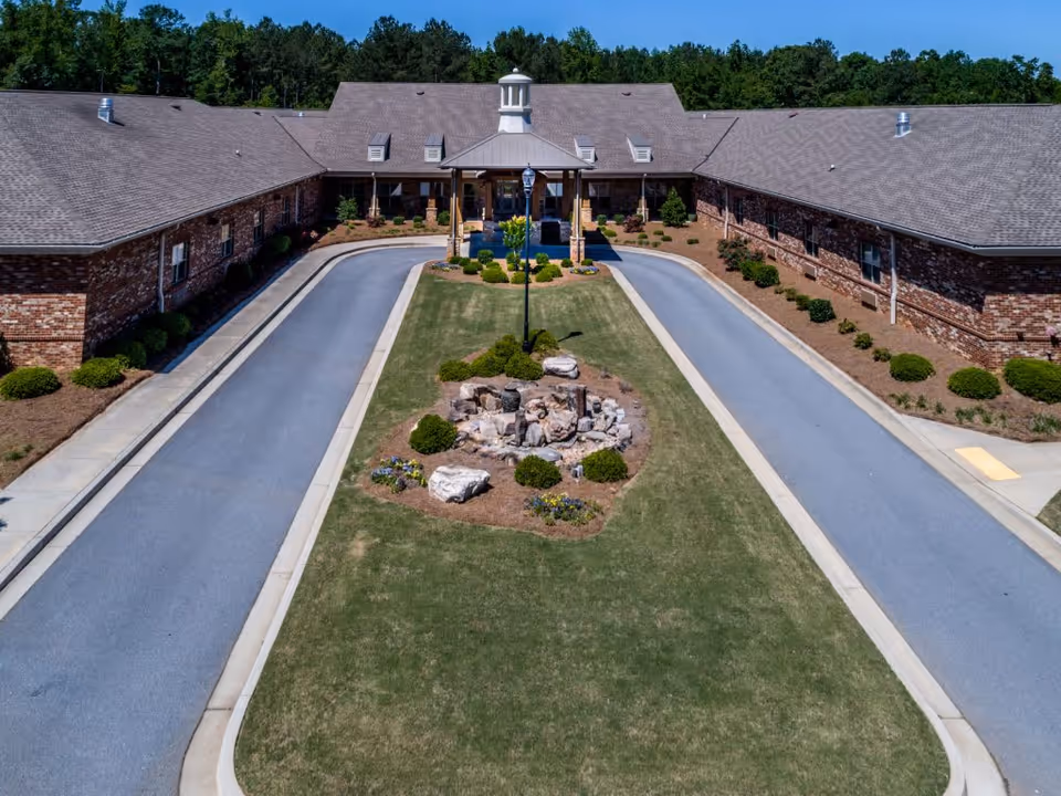 Aerial view of the entrance driveway to a single-story brick building with a central covered portico. The driveway splits into two lanes around a landscaped grassy median with rocks, shrubs, and a lamppost. Trees are visible in the background.