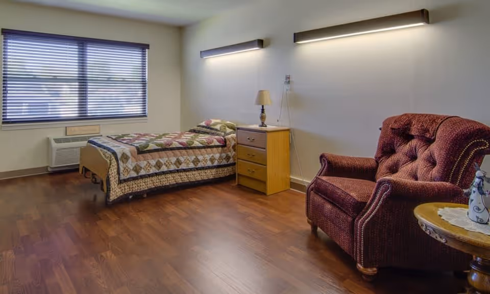 A nursing facility bedroom with a single bed covered by a quilt, a wooden nightstand, and a red upholstered armchair.