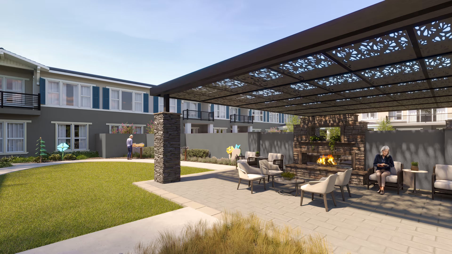 Outdoor seating area at The Ivy At Blue Oaks featuring a covered patio with decorative panels, stone pillars, a stone fireplace with a fire burning, and several cushioned chairs and tables. In the background, there is a green lawn, a walking path, and a person standing near a wall with colorful decorations. The building exterior with windows and balconies is visible behind the wall.