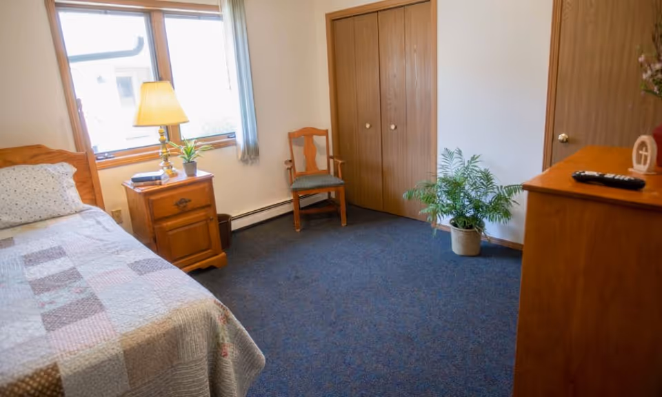 A cozy bedroom with a single bed covered in a patchwork quilt, a wooden nightstand with a lamp and a small plant, a wooden chair, a potted plant on the floor, and a wooden dresser with a remote control on top. There is a window with light blue curtains and a double-door closet with wooden doors.