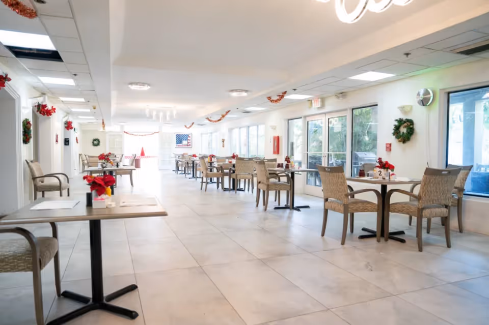 Bright, spacious dining room with multiple small tables and chairs arranged along windows, decorated for the holidays.