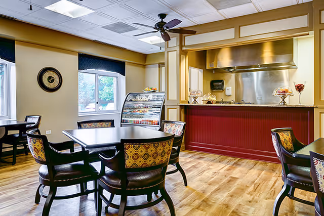 Dining area with round tables and upholstered chairs, a service counter with stainless hood, and a refrigerated display case by windows.