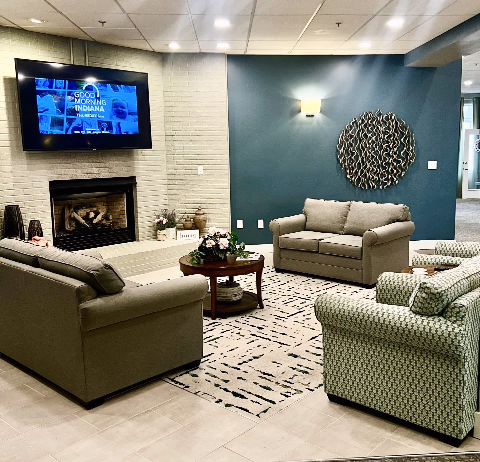 A cozy living room area with two beige sofas and two patterned armchairs arranged around a wooden coffee table with a floral centerpiece. A flat-screen TV is mounted above a white brick fireplace on the left wall. The wall behind one sofa is painted teal and decorated with a round metal wall art piece and a wall sconce light. The floor is tiled with a patterned area rug in the seating area.