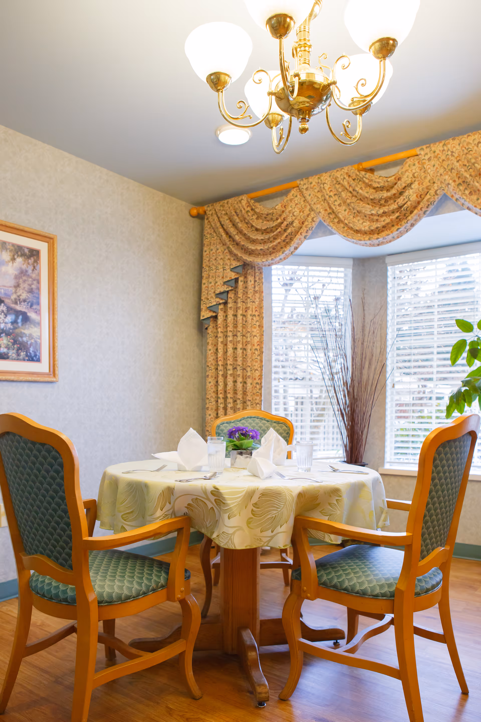 A small dining area with a round table covered with a floral tablecloth, set with napkins, glasses, and a small flower centerpiece. Four wooden chairs with green cushioned seats surround the table. A window with white blinds and floral drapes is in the background, along with a decorative vase with tall branches. A chandelier with five lights hangs from the ceiling.