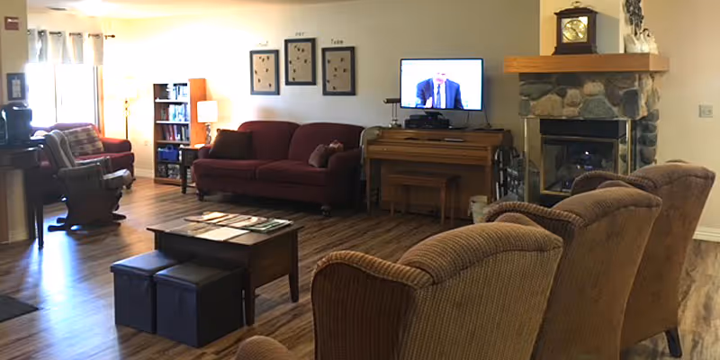 A cozy senior living communal lounge with sofas, armchairs, a TV on a wooden piano, a stone fireplace, and wood flooring.