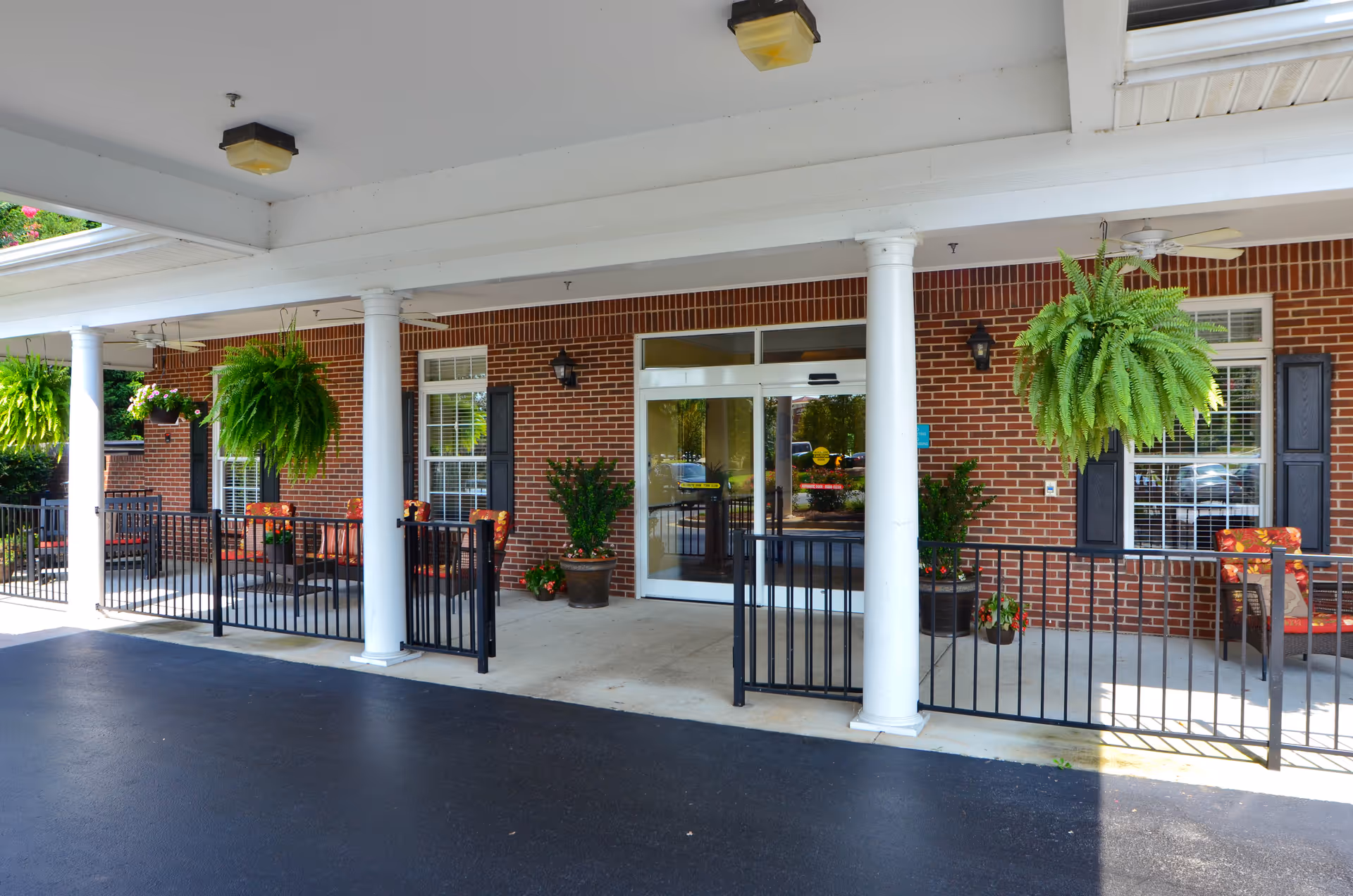 Covered entrance of a brick senior living facility with white columns, hanging ferns, outdoor seating, and automatic glass doors.