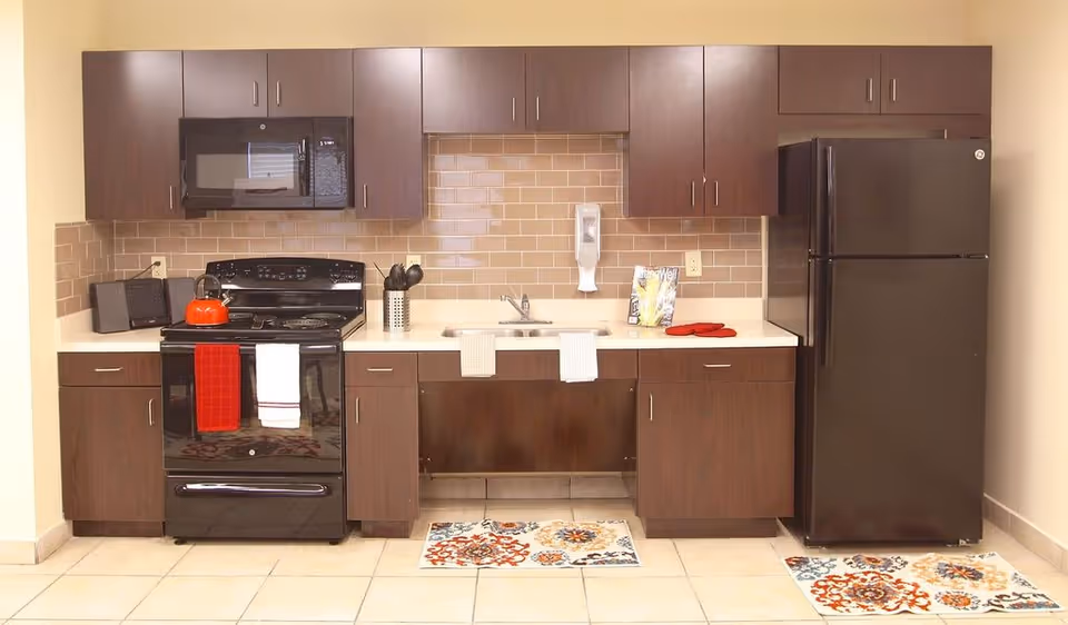 A kitchen area with dark brown cabinets, a black microwave, stove with a red kettle and two towels hanging on the oven door, a sink with two towels hanging on the cabinet handles, and a black refrigerator. The backsplash is made of beige subway tiles, and there are two colorful patterned rugs on the tiled floor.