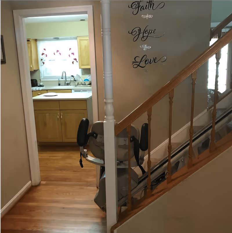 View of a hallway leading to a kitchen with wooden cabinets and a window with floral curtains. On the right side, there is a staircase with a stairlift installed. The wall next to the staircase has decorative words 'Faith', 'Hope', and 'Love' with small butterfly accents.