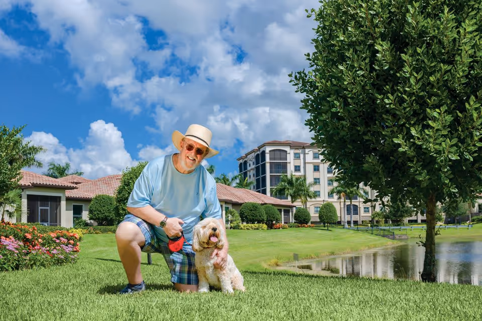 An elderly man wearing a hat, sunglasses, a light blue shirt, and plaid shorts is kneeling on green grass next to a small fluffy dog. They are outdoors on a sunny day with a blue sky and white clouds. Behind them are well-maintained gardens, a pond, trees, and residential buildings with tiled roofs.