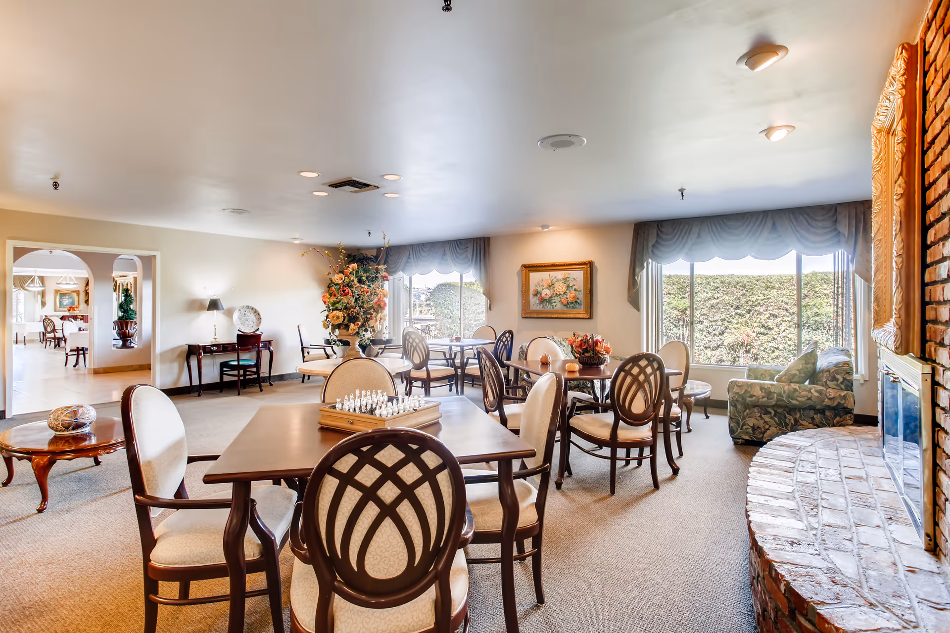 A bright and spacious common area in a senior living facility with multiple round tables and chairs arranged for socializing or activities. There is a chessboard on one table, a large floral arrangement on another, and a cozy seating area with a floral-patterned couch near a brick fireplace. Large windows with drapes let in natural light, and a painting hangs on the wall. The room has carpeted floors and soft ceiling lighting.