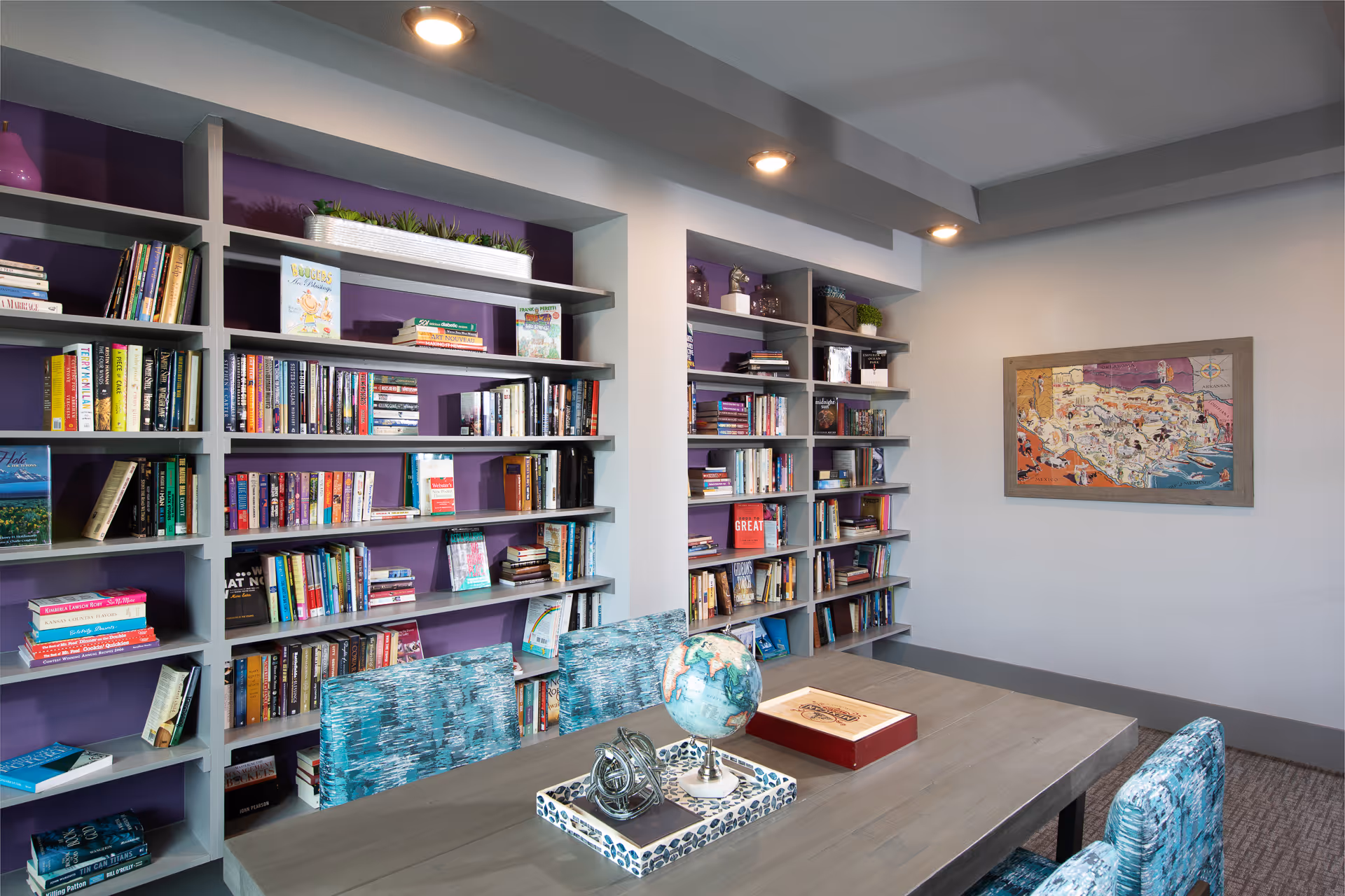A bright reading room with wall bookshelves, a table topped with a globe and decorative tray, and patterned upholstered chairs.