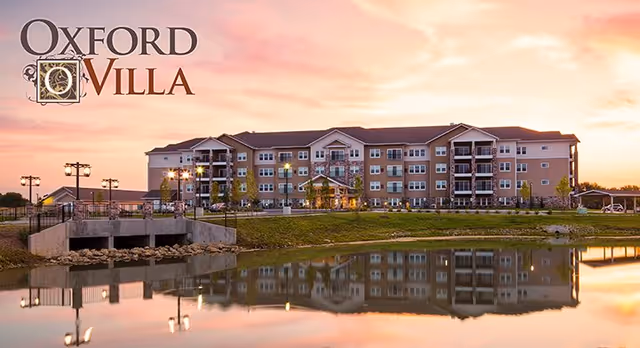 Exterior view of Oxford Villa Active Senior Apartments at sunset, showing a multi-story building reflected in a calm body of water in the foreground.