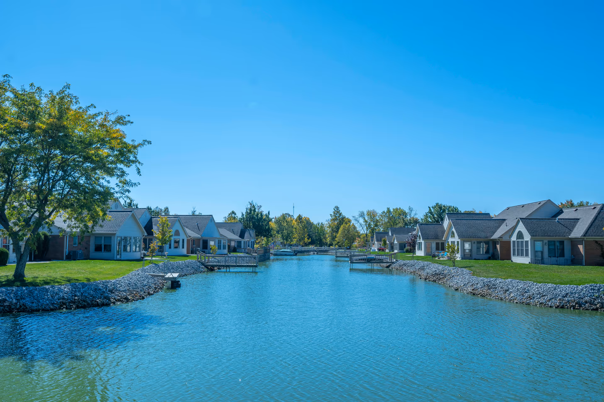 A serene waterway lined with small residential buildings on both sides under a clear blue sky. The buildings have sloped roofs and large windows, with green lawns and trees along the water's edge.