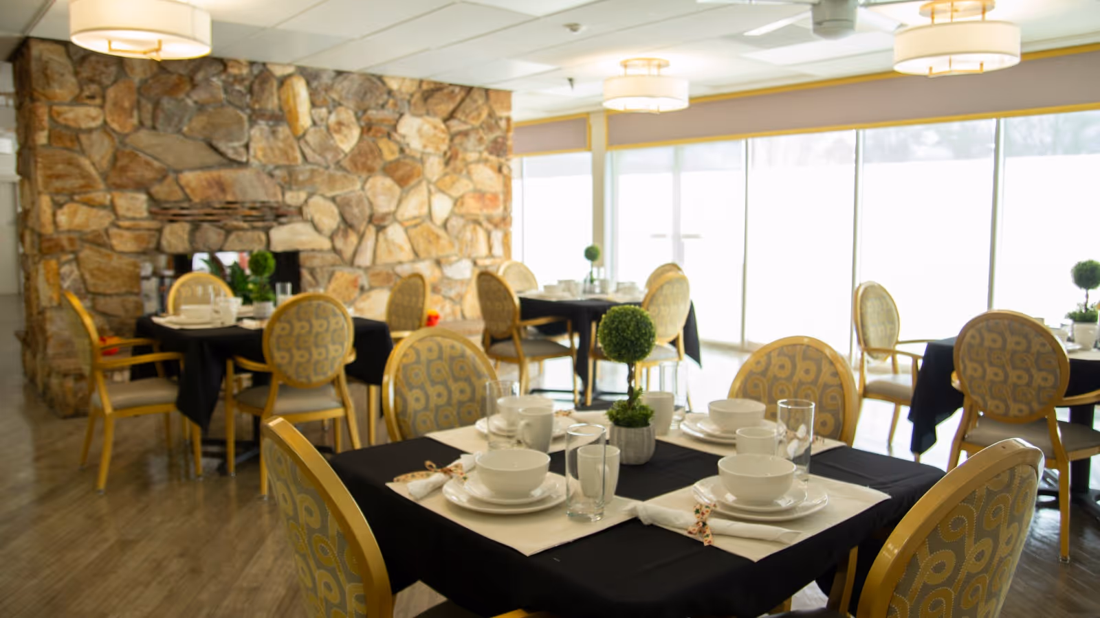 A dining room with multiple tables set with white dishes, bowls, glasses, and napkins on black tablecloths. The room features patterned chairs, a stone fireplace on one side, and large windows allowing natural light to fill the space.
