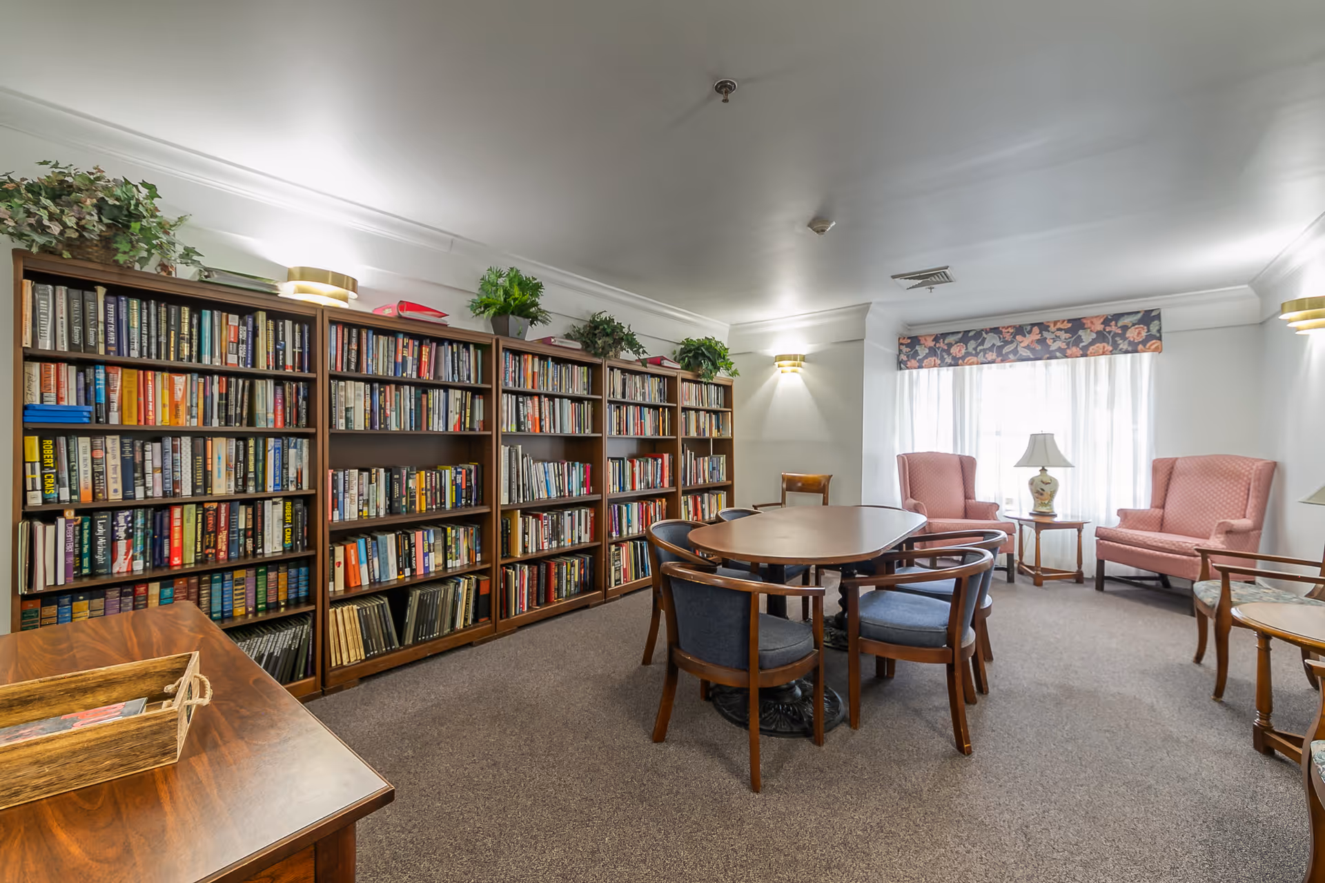 A cozy reading room with a large wooden bookshelf filled with books along one wall, a round wooden table surrounded by six chairs in the center, and two pink upholstered armchairs near a window with sheer white curtains and a floral valance. There are also small side tables with lamps and some potted plants on top of the bookshelf.