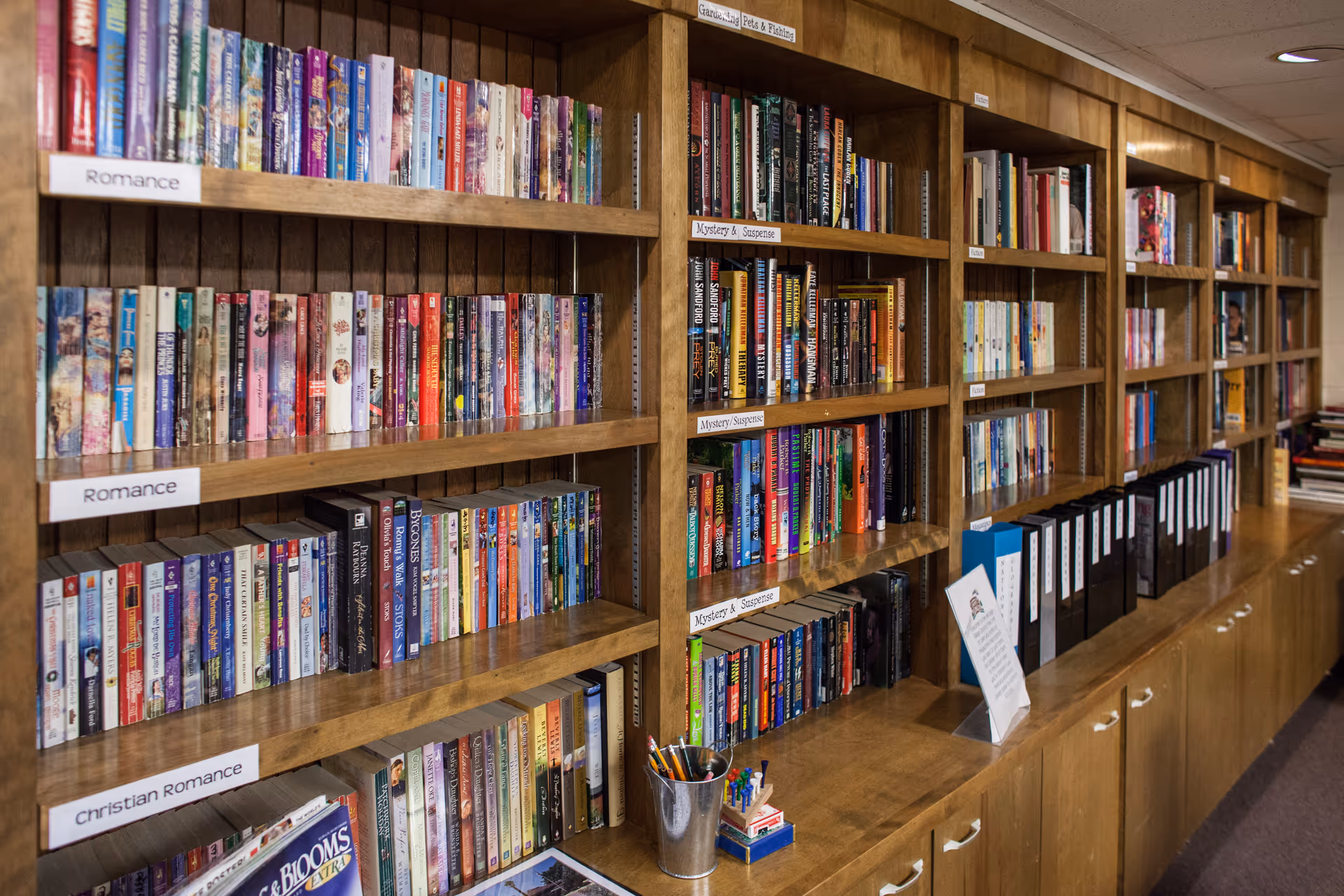 Wooden bookshelves filled with various books organized by genres such as Romance, Christian Romance, Mystery & Suspense, and Gardening, Pets & Fishing. The shelves also hold binders and a small container with pens and pencils. The setting appears to be a library or reading area within a facility.