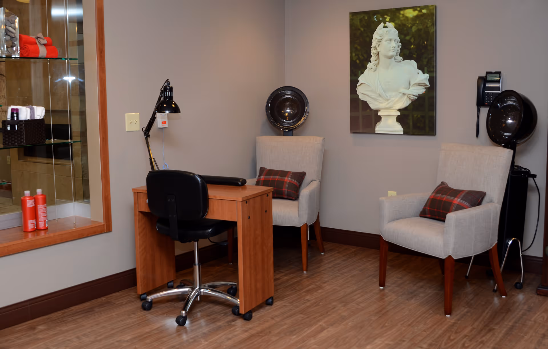 A cozy salon area with two beige armchairs each with a plaid cushion, two black hair dryer chairs, a wooden desk with a black swivel chair, a desk lamp, and a wall-mounted phone. A framed bust sculpture is hung on the wall above the armchairs. Shelves with towels and hair products are visible through a glass window on the left.