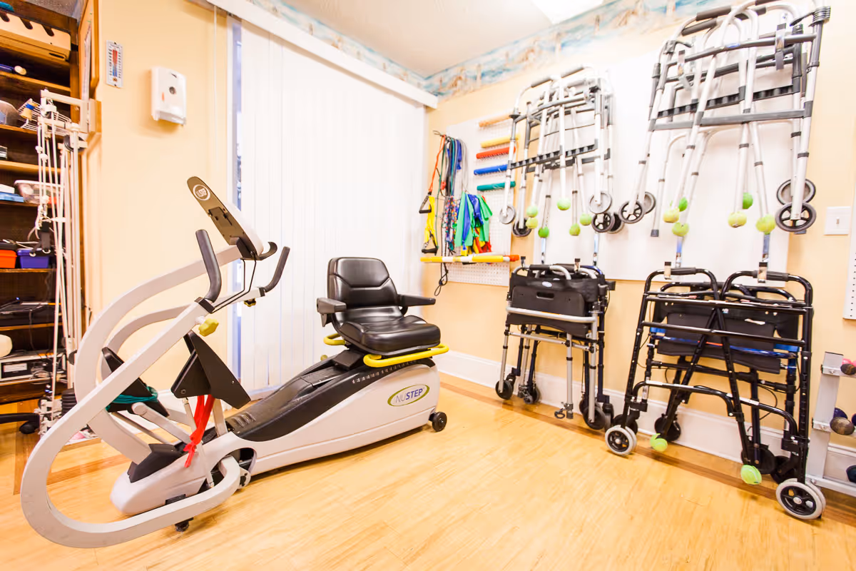 A rehabilitation and wellness room featuring a NuStep exercise machine with a black seat, several walkers with tennis balls on their legs hanging on the wall, and various resistance bands and exercise equipment organized on pegboards. The room has light-colored walls and a wooden floor.