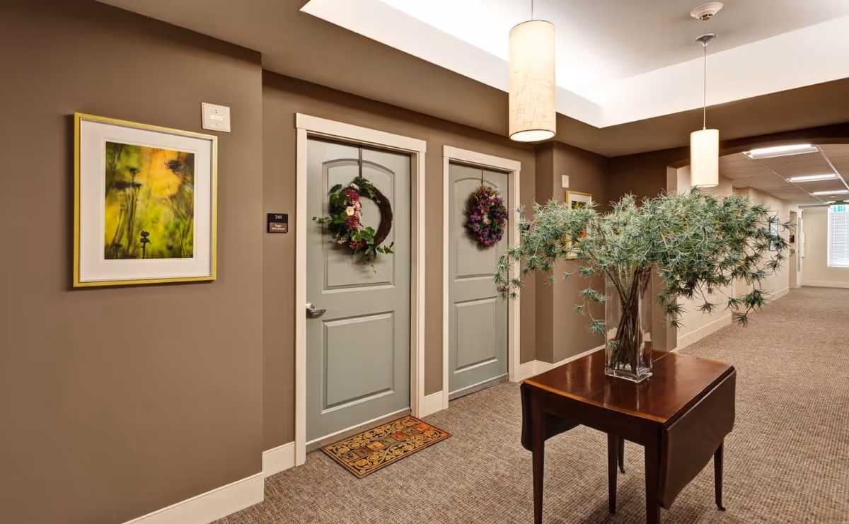 A hallway in an assisted living facility with two light green doors decorated with floral wreaths. A small table with a glass vase holding green foliage is positioned in the hallway. The walls are painted brown and adorned with framed artwork. The floor is carpeted, and pendant lights hang from the ceiling.