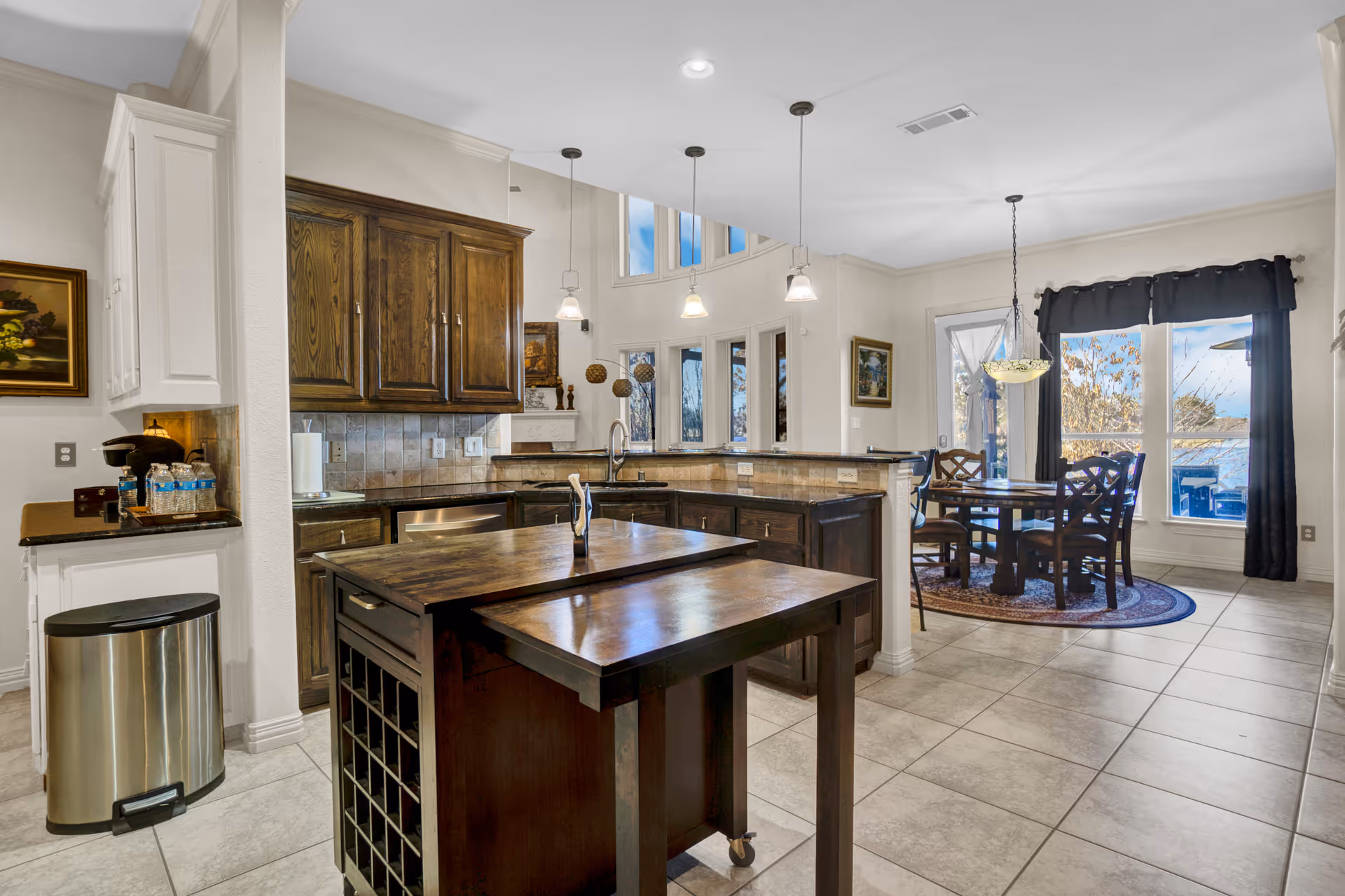 A spacious kitchen with dark wood cabinets and a central island with a pull-out table. The kitchen has tiled floors and a tiled backsplash. Pendant lights hang from the ceiling. In the background, there is a dining area with a round wooden table and four chairs, a large window with black curtains, and a door leading outside.