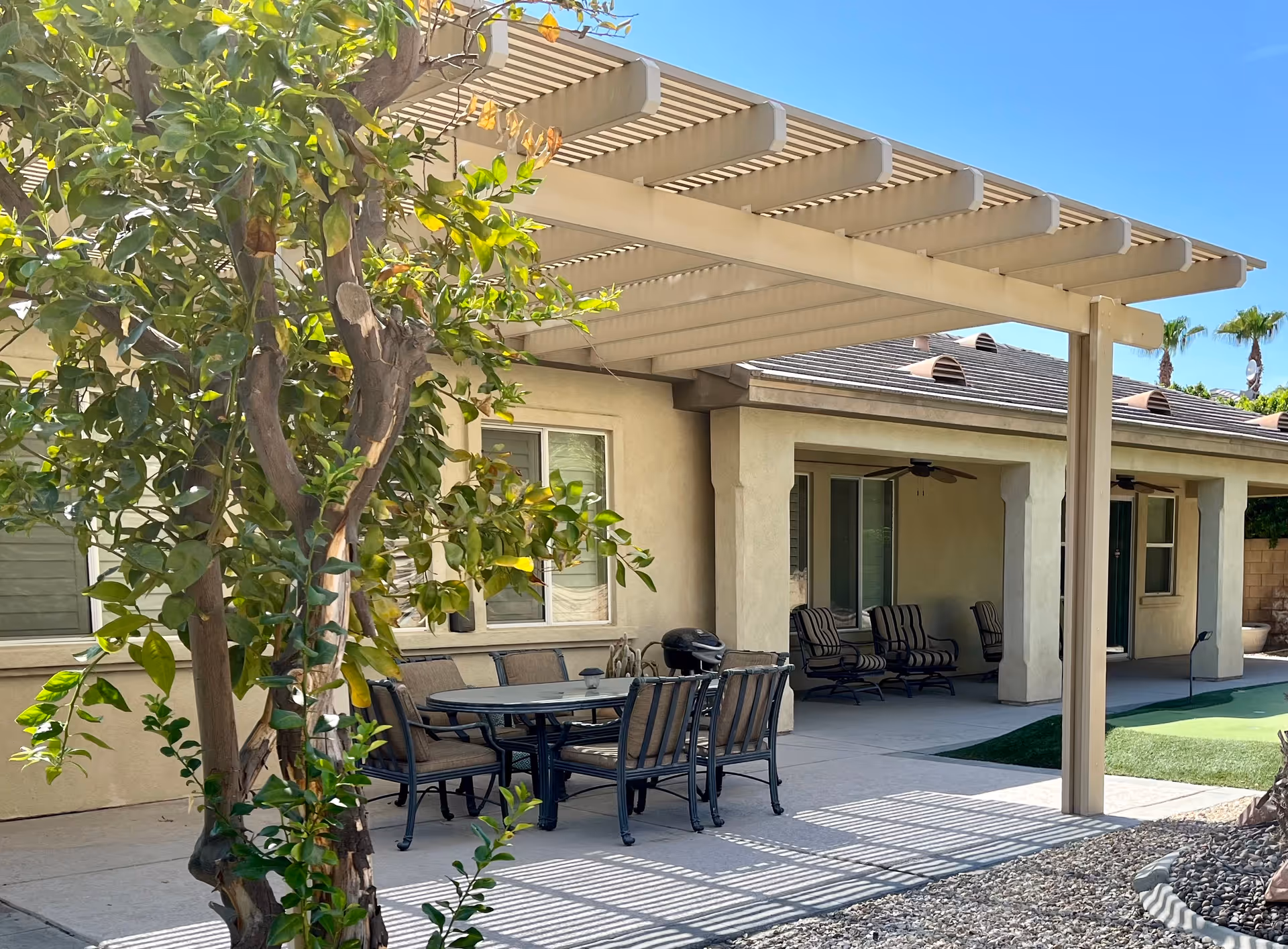 Outdoor patio area at Shiloh Senior Living in La Quinta featuring a shaded pergola with a dining table and chairs, a tree in the foreground, and additional seating under a covered porch attached to a beige building with windows and ceiling fans.