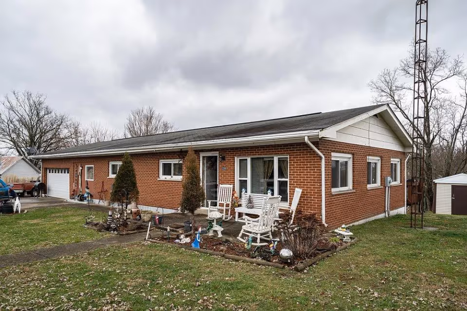 Exterior view of a single-story brick building with a small garden and white rocking chairs on a concrete patio. The sky is overcast and there is a metal tower structure on the right side of the building.