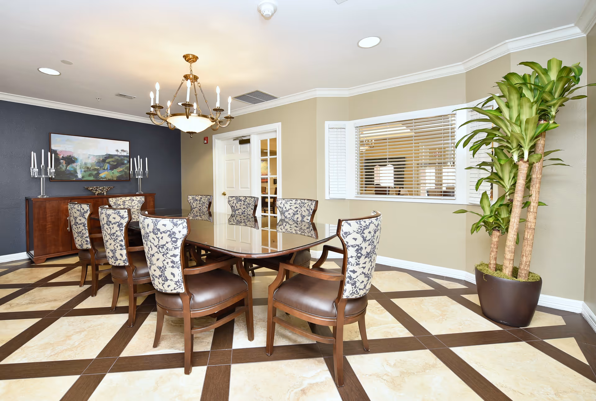 A bright dining room featuring a glass-topped table surrounded by patterned upholstered chairs, a chandelier, a sideboard, and a large potted plant.