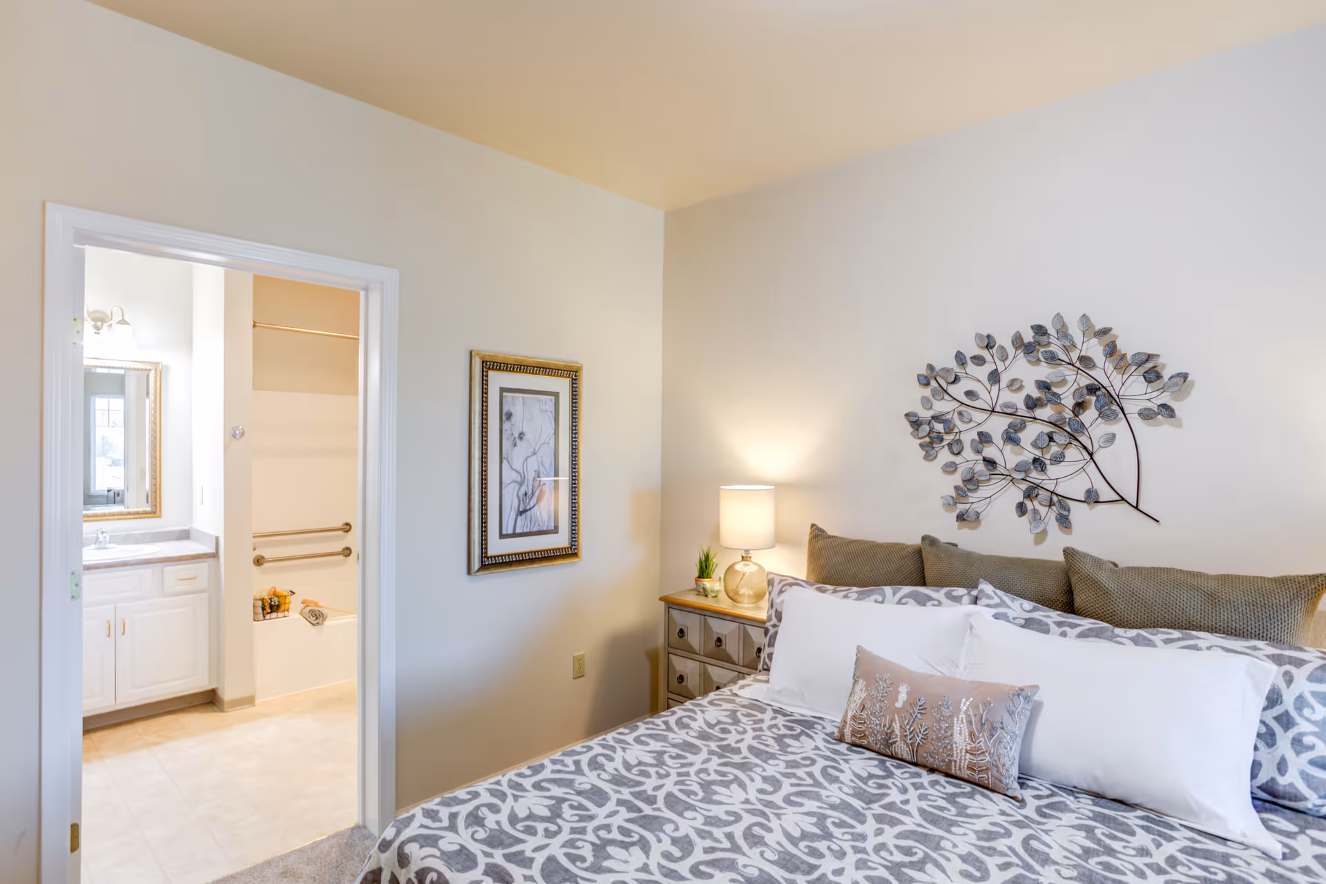 A bedroom with a bed featuring patterned bedding and multiple pillows, a nightstand with a lamp and small plant, a decorative metal wall art shaped like a tree branch above the bed, and an open door leading to a bathroom with a bathtub and vanity.