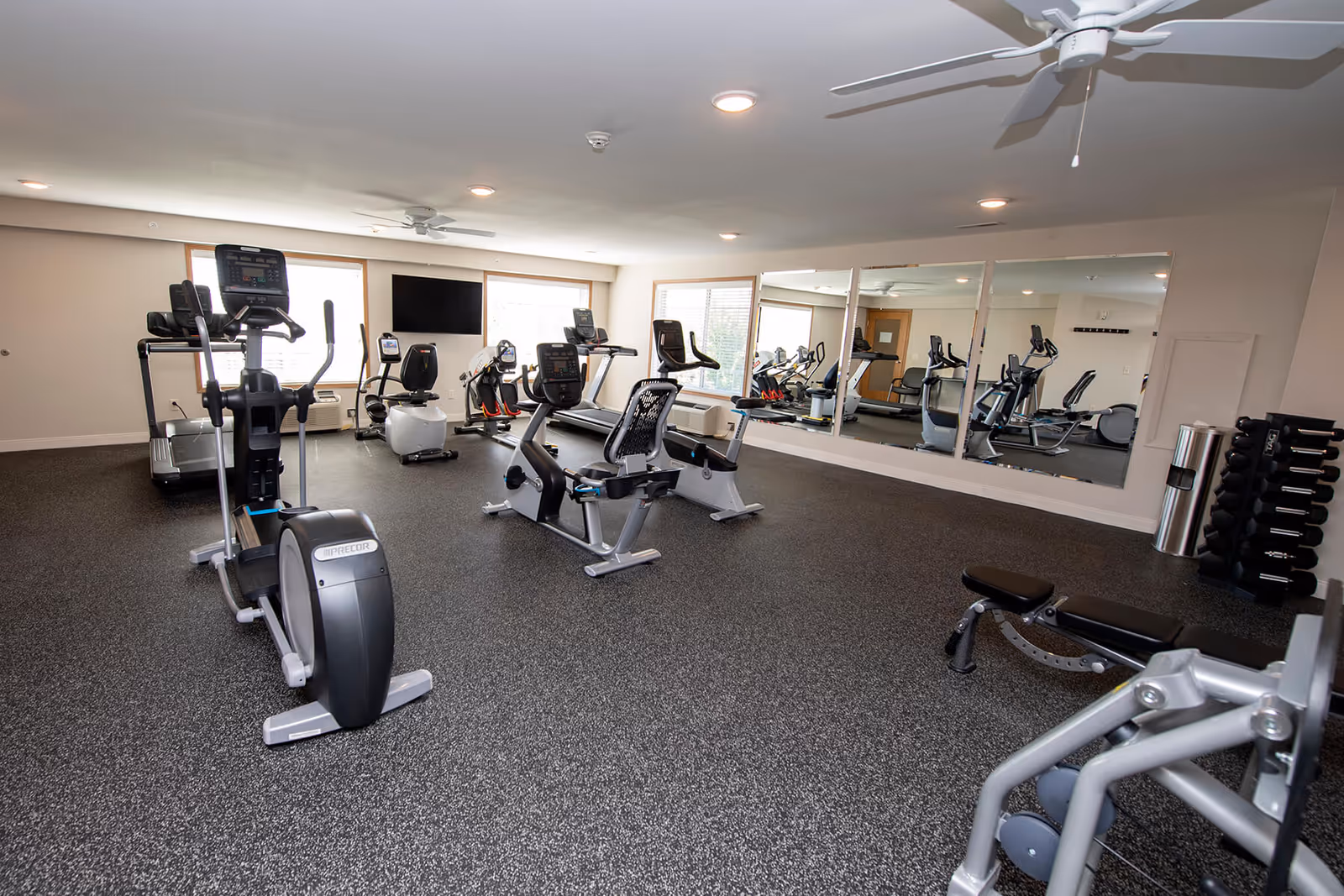 A fitness room with various exercise equipment including stationary bikes, elliptical machines, and weight benches. The room has large windows letting in natural light, a wall-mounted TV, and a large mirror covering one wall. The floor is covered with rubber gym flooring.