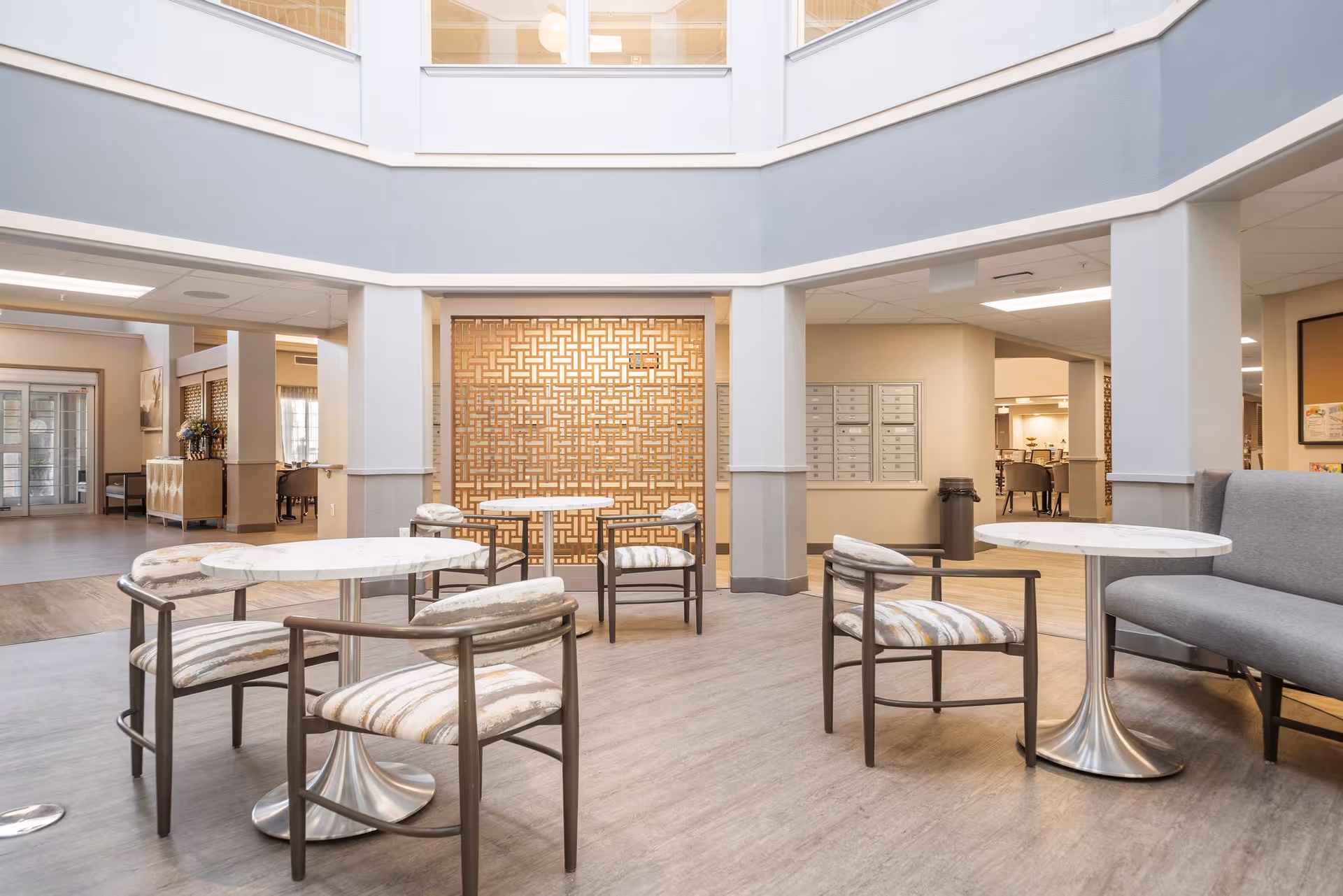 A bright and spacious interior common area with modern round tables and cushioned chairs arranged on a wood-patterned floor. The space features a decorative wooden lattice wall, mailboxes on the wall, and a gray cushioned bench. The area is well-lit with overhead lighting and has an open layout with visible adjoining rooms.