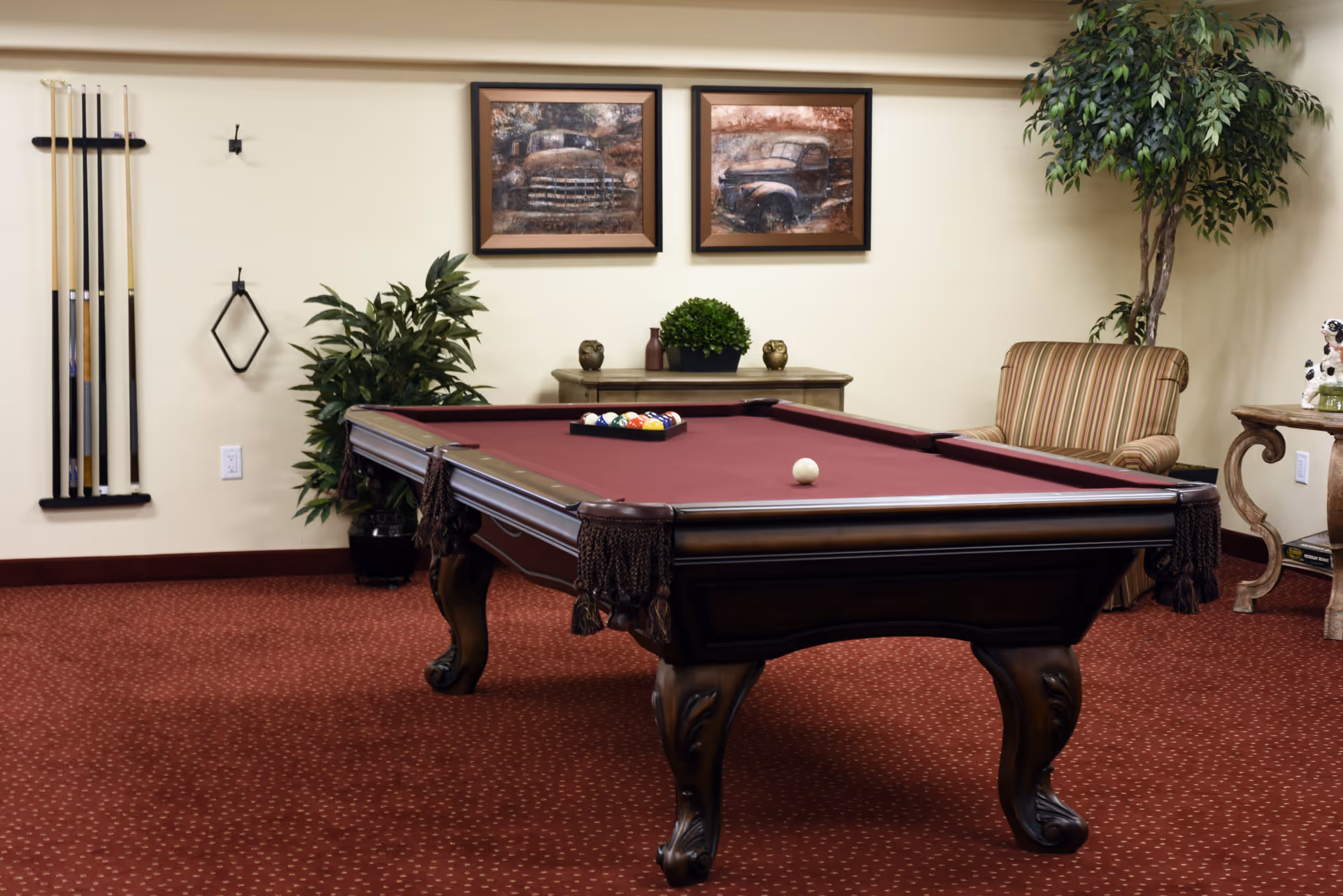 A carpeted recreation room featuring a maroon pool table, wall-mounted cues, framed artwork, plants and upholstered seating.